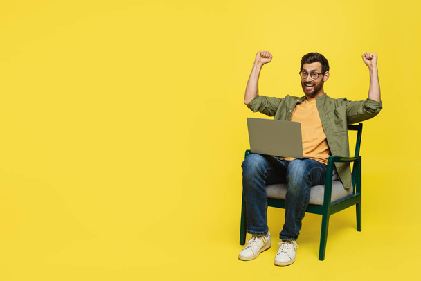 Overjoyed man sitting on chair with laptop and shaking fists, celebrating victory and success over yellow studio background, copy space. Great news concept - Foto, imagen