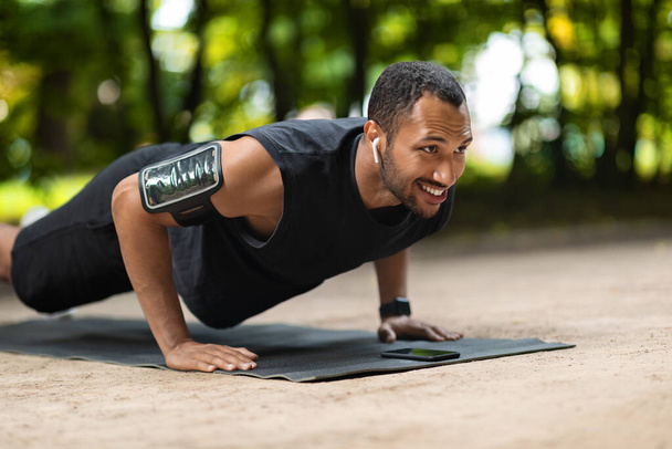 Κίνητρο όμορφος νεαρός αφροαμερικανός τύπος αθλητής planking στο δημόσιο πάρκο, άσκηση στο στρώμα γυμναστικής, κοιτάζοντας χώρο αντίγραφο και χαμογελώντας, χρησιμοποιώντας ακουστικά και περιβραχιόνιο smartphone - Φωτογραφία, εικόνα