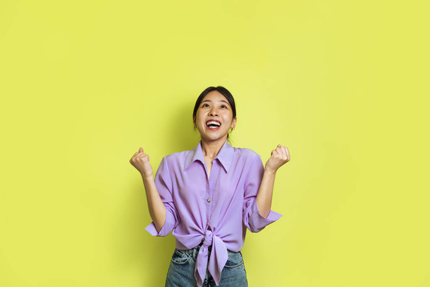 Joyful Asian Lady Gesturing Yes And Shaking Fists With Both Hands Looking Up Standing Over Yellow Background. Woman Celebrating Victory And Great Success Posing In Studio - Foto, Bild