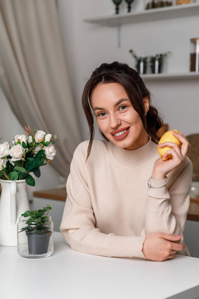 mooi jong brunette meisje in de keuken in een trui met citroen. mooie glimlach. - Foto, afbeelding