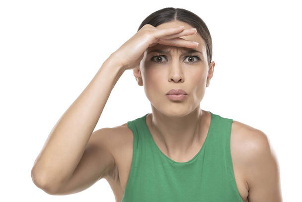 Περίεργη μελαχρινή γυναίκα με πράσινο πουκάμισο κοιτάζει μακριά με το χέρι πάνω από το κεφάλι, προσπαθώντας να δει κάτι - Φωτογραφία, εικόνα