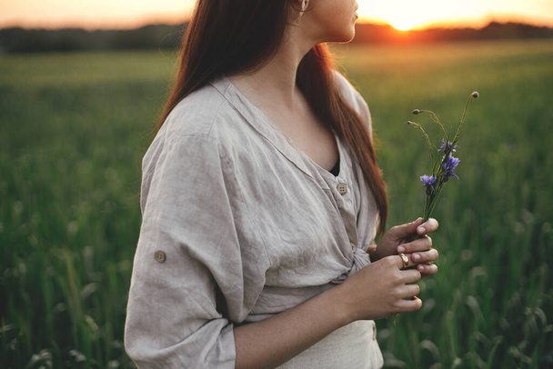 Woman holding wildflowers in wheat field in warm sunset light. Tranquil atmospheric moment. Close up of young female in rustic dress with flowers in hands in evening summer countryside. - Photo, image