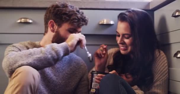 Fun, food and young couple eating ice cream in a kitchen at home, bonding, playful and looking happy. man and woman being affectionate and flirting, dessert and relax in the kitchen floor together. - Filmmaterial, Video