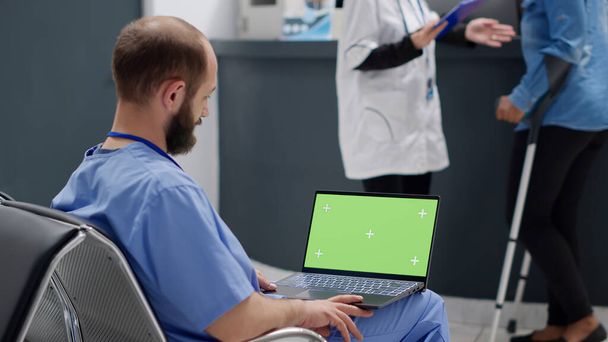 Asystent medyczny pracujący na laptopie z zielonym ekranem w holu recepcyjnym, siedzący w poczekalni w klinice. Korzystanie z izolowanego wyświetlacza makietowego z pustym szablonem klawisza chroma i tłem copyspace. - Zdjęcie, obraz