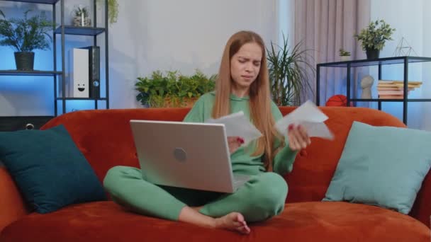 Stressed girl at home room looking at unpaid bank debt bills, doing paperwork, planning budget, calculate finances mortgage payments. Displeased young child kid throws paper bills sitting on couch - Séquence, vidéo