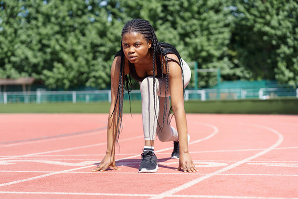 Black young sportive female athlete prepares low start running action on track of city stadium against green trees in park. African American woman with long braids trains before Olympics on sunny day - Φωτογραφία, εικόνα