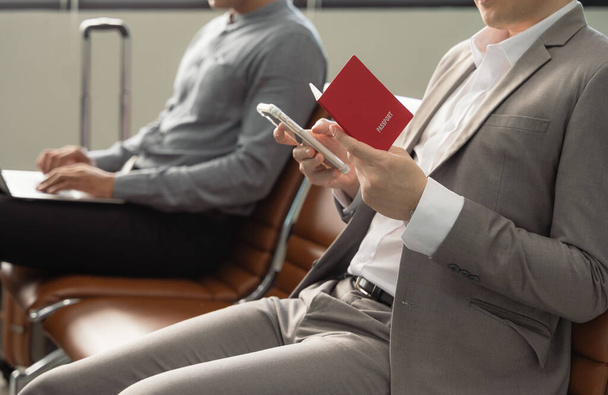 Businessmen waiting for flight in airport terminal. Man in suit holding passport sitting and working on mobile phone while other guy working on laptop. Business travel concept. - Photo, Image