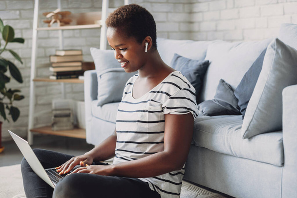 Молодая счастливая афроамериканка, сидящая на полу и наслаждающаяся работой дома на ноутбуке. Просмотр Интернета, проведение времени дома в гостиной. Студент - Фото, изображение