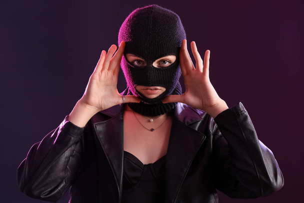 Young woman in balaclava and leather jacket with hands near face against dark background - Valokuva, kuva