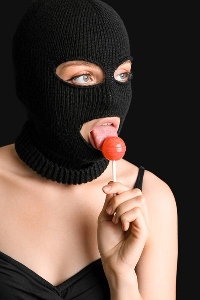 Young woman in balaclava licking lollipop on black background - Photo, image