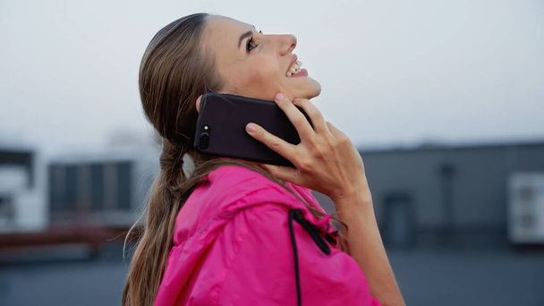 Side view of smiling sportswoman talking on smartphone on roof in evening  - Fotografie, Obrázek