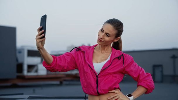 Smiling sportswoman taking selfie on smartphone on roof in evening  - Zdjęcie, obraz
