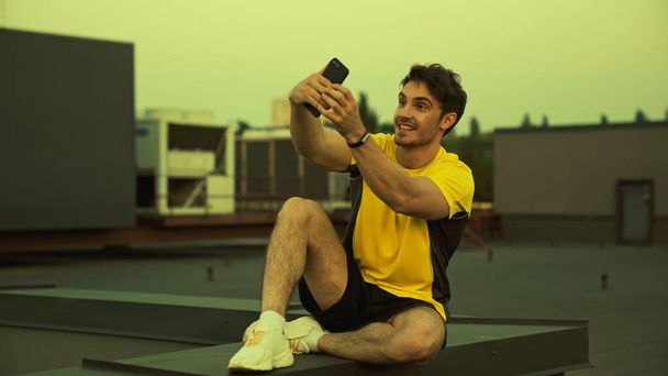 Smiling sportsman in sportswear and sneakers taking selfie on roof in evening  - Fotoğraf, Görsel