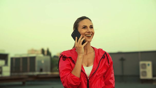 Smiling sportswoman talking on mobile phone on roof in evening  - Фото, изображение