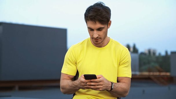 Sportsman in yellow sportswear using mobile phone on blurred roof in evening  - Fotografie, Obrázek
