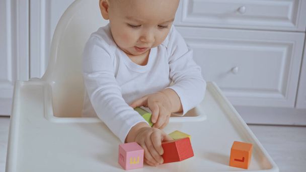 focused baby girl sitting in feeding chair and playing with wooden cubes - Foto, afbeelding