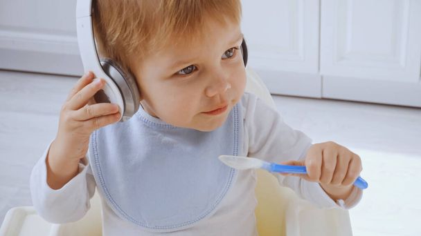 cute toddler boy listening music in headphones while holding plastic spoon - Foto, afbeelding