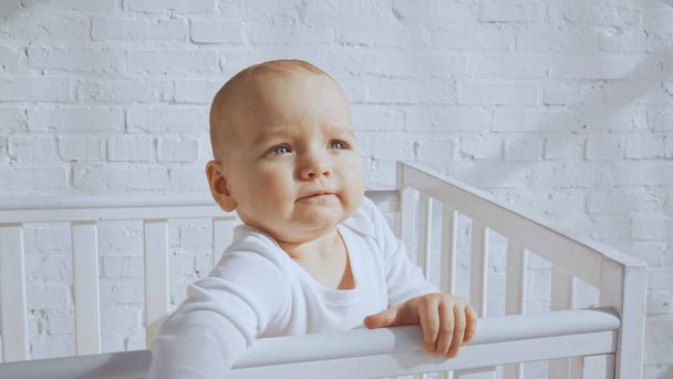 чарівна дитина з блакитними очима, що стоїть у білому дитячому ліжечку вдома
  - Фото, зображення