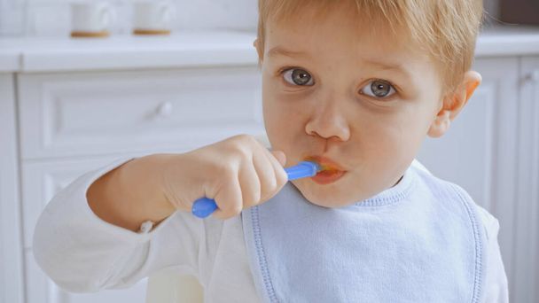 toddler boy with grey eyes holding spoon and eating carrot puree  - Foto, afbeelding