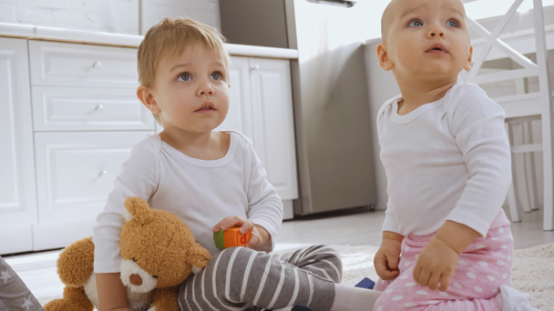 toddler boy sitting on carpet with teddy bear near baby sister and looking away - Photo, image