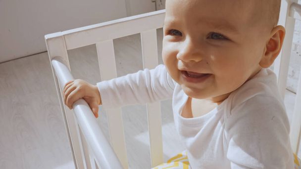 close up of cheerful baby girl standing in white baby crib at home   - Фото, изображение
