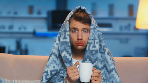 sick man with blue eyes holding cup of tea while sitting under blanket in living room - Fotó, kép