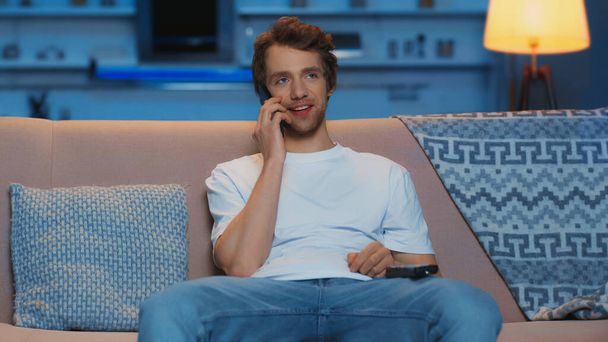 happy young man holding remote controller while talking on smartphone in living room  - Foto, Imagem
