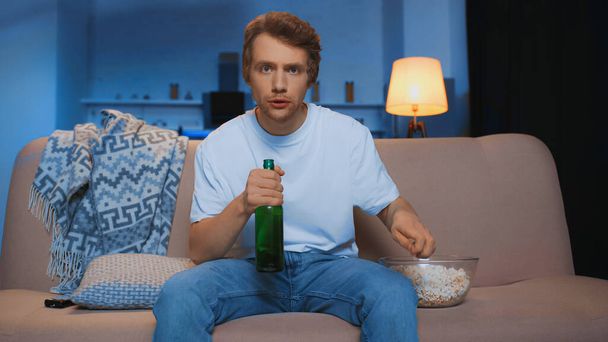 молодий чоловік з пляшкою пива сидить на дивані біля миски попкорну під час перегляду чемпіонату
   - Фото, зображення