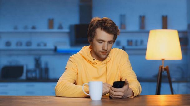 зосереджений молодий чоловік використовує смартфон біля чашки чаю на дерев'яному столі
 - Фото, зображення