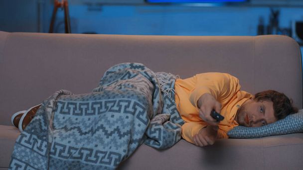 young man in yellow hoodie lying under blanket and watching tv on sofa at night  - Foto, afbeelding