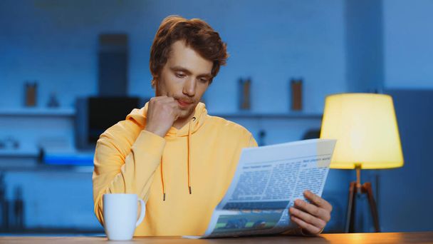 young man in yellow hoodie reading newspaper near cup of tea at home  - Foto, Imagem