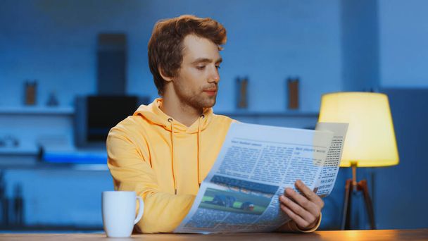 young man in yellow hoodie reading newspaper near cup with beverage at home - Photo, image