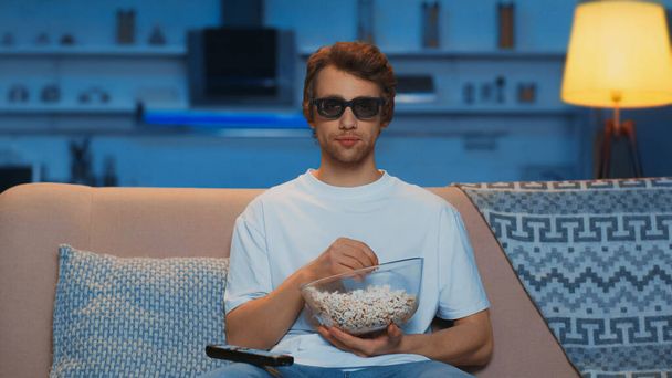 young man in 3d glasses holding bowl with popcorn while watching movie in living room  - Zdjęcie, obraz