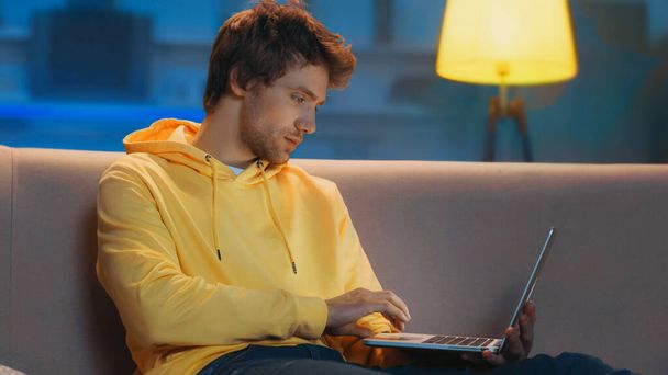 young freelancer in yellow hoodie using laptop while working from home in evening  - Фото, изображение