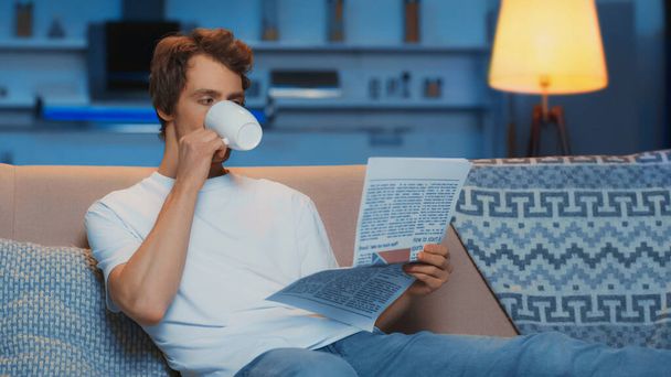 young man in white t-shirt reading newspaper and drinking tea while resting on couch - Fotoğraf, Görsel