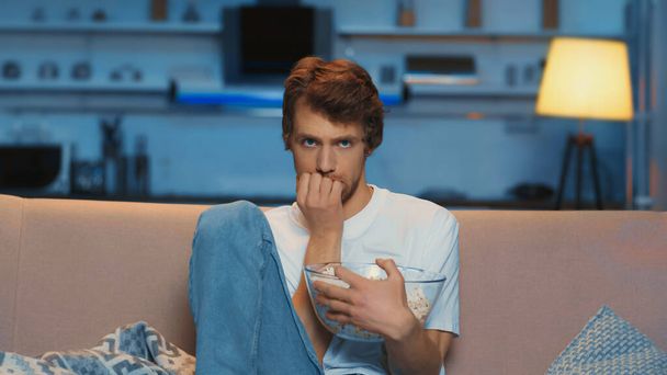 tensed young man holding bowl of popcorn while watching scary movie  - Photo, Image