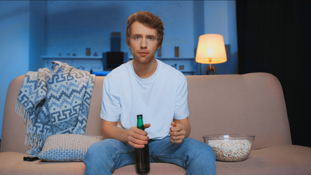 focused man with bottle of beer sitting on couch near bowl of salty popcorn while watching championship  - Фото, изображение