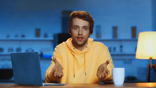 young man in yellow hoodie talking while gesturing near laptop and cup of coffee in evening  - Fotografie, Obrázek