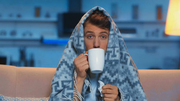 sick man with blue eyes drinking hot beverage while sitting under blanket in living room  - 写真・画像