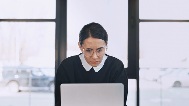 serious and thoughtful businesswoman in eyeglasses looking at laptop in office - 写真・画像