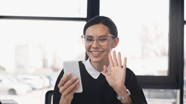 cheerful businesswoman in eyeglasses waving hand during video call on smartphone - Фото, изображение