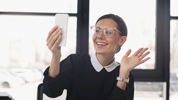 excited businesswoman in eyeglasses waving hand at mobile phone during video chat - Foto, Bild