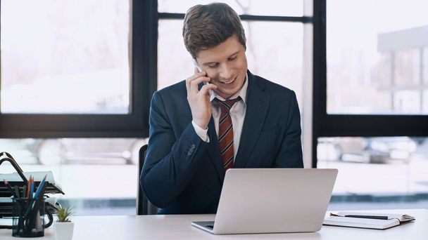 happy businessman talking on smartphone near laptop and notebook in office - Zdjęcie, obraz