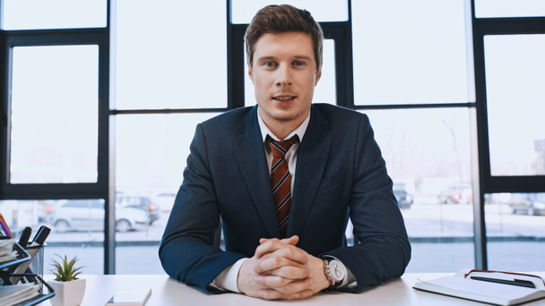 positive businessman sitting at workplace with clenched hands and looking at camera - Foto, afbeelding
