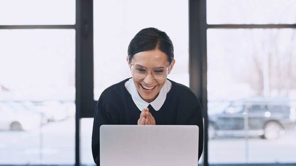 joyful businesswoman in eyeglasses showing wow gesture with folded hands near laptop - 写真・画像
