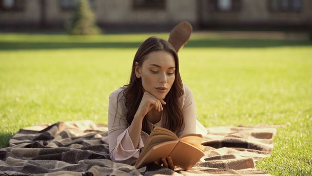 Brunette woman reading book while relaxing on blanket in park  - Fotó, kép