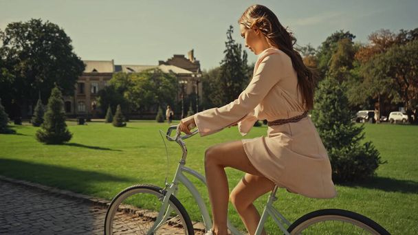 Side view of brunette woman in dress cycling in summer park  - Zdjęcie, obraz