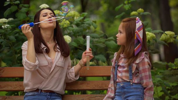 Woman blowing soap bubbles near redhead daughter on bench in park  - Photo, image