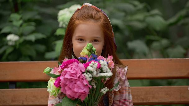 Red haired kid looking at camera near flowers on bench in park  - 写真・画像