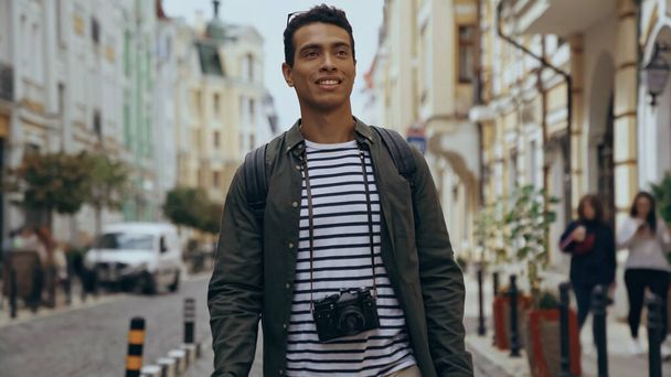 Smiling bi-racial tourist with film camera walking on urban european street  - Foto, imagen